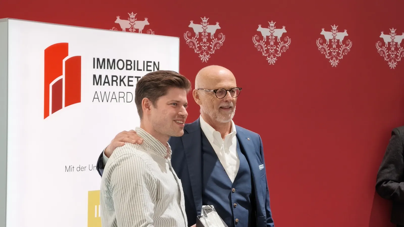 Zwei Männer lächeln bei der Übergabe der gläsernen Trophäe vor dem Banner des Immobilien-Marketing-Award und einer roten Wand mit weissen Ornamenten.