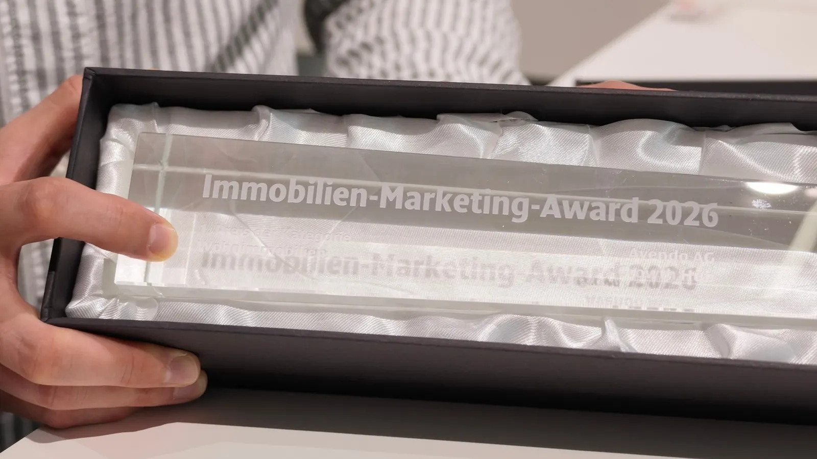 Gläserne Trophäe mit der Gravur «Immobilien-Marketing-Award 2026» in einer mit weissem Satin ausgeschlagenen Schachtel, von zwei Händen gehalten